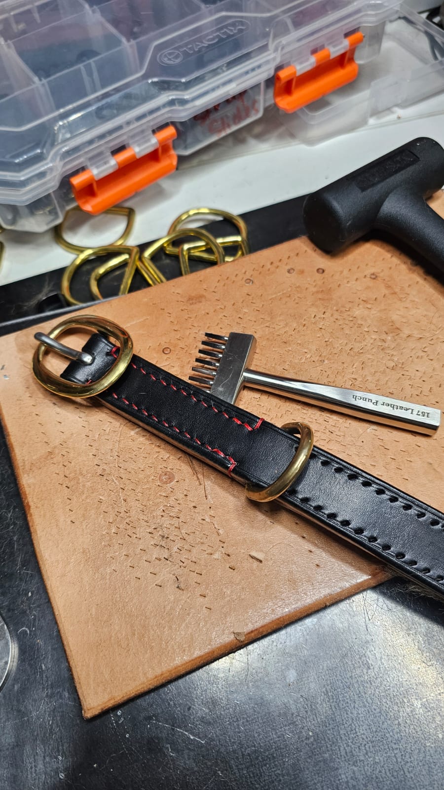 Black leather strap with gold buckle on a wooden surface, with tools and a clear plastic container in the background.