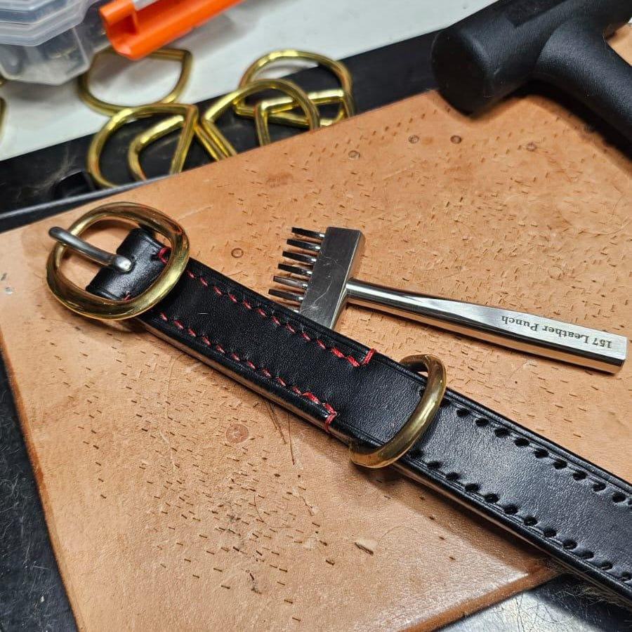 Black leather strap with gold buckle on a wooden surface, with tools and a clear plastic container in the background.